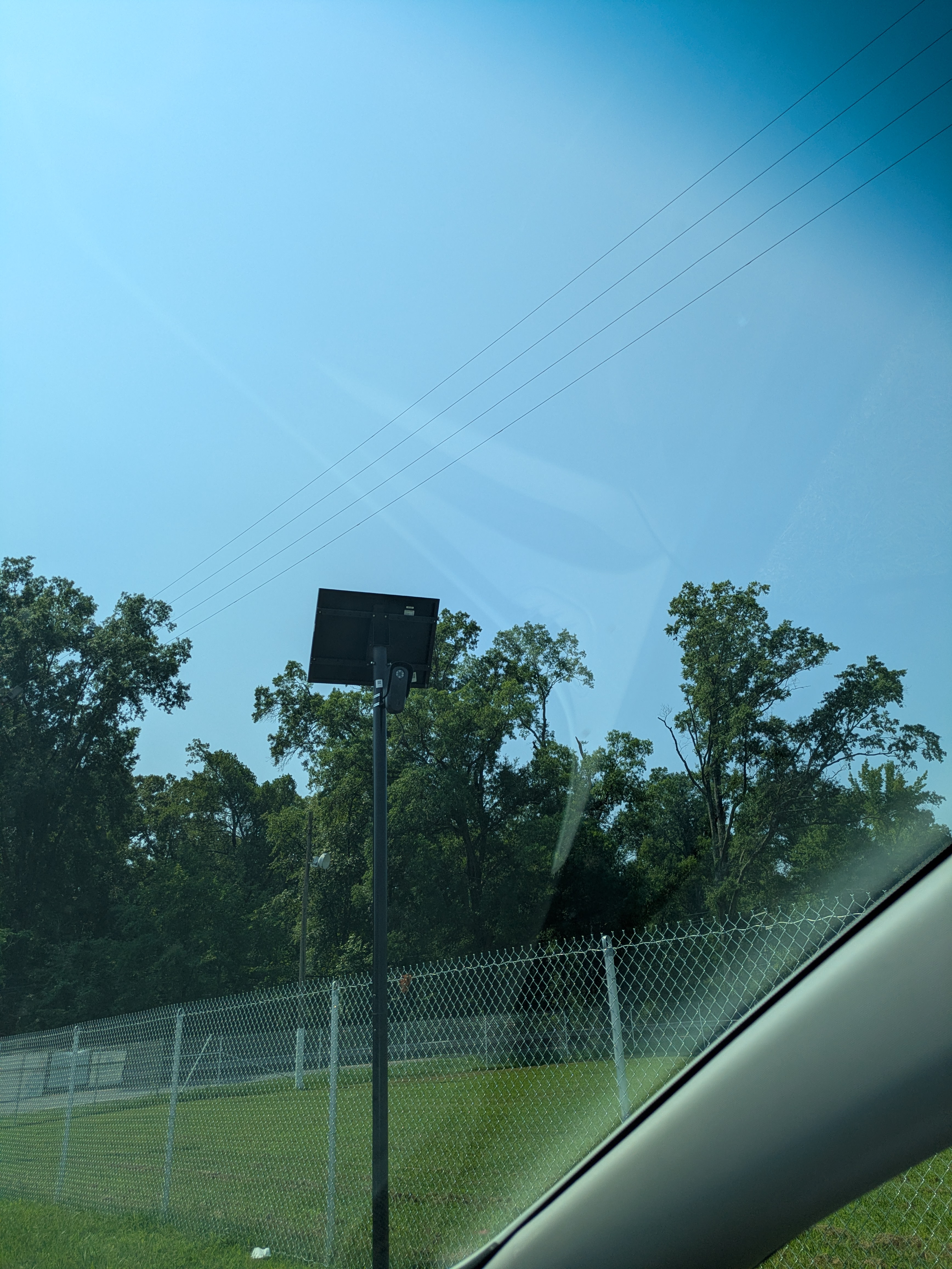 A Flock camera and its solar panel mounted on a black pole, standing behind a chain-link fence at the edge of a grass field, photographed from a car.